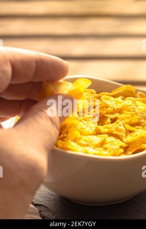 Selective focus of a person picking up cornflakes for breakfast with his hand from a full bowl on a wooden table. Stock Photo