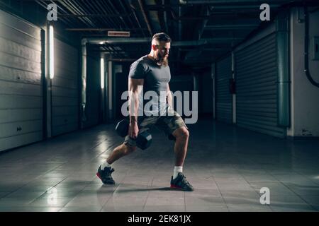 Side view of muscular attractive caucasian bearded man doing lunges with dumbbells in underground hallway. Storage units and pipes in background. Stock Photo