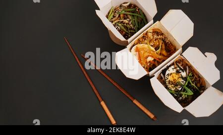 Noodles with pork and vegetables in take-out box on black table. Asian food delivery. Food in paper containers on black background Stock Photo