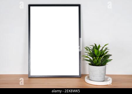 Black wooden vertical frame with white blank card and green plant in concrete pot on wooden table on gray wall background. Mockup, template for your d Stock Photo