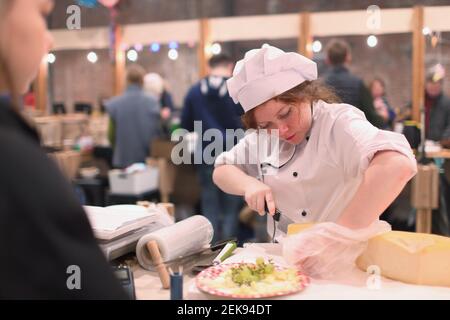 Female market vendor cutting cheese during the fair in Sevkabel public space, St. Petersburg, Russia Stock Photo