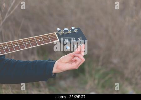 A young white man, musician and artist, tune the strings of an elegant black guitar, with the pegs, in the countryside, on a cloudy afternoon. Stock Photo