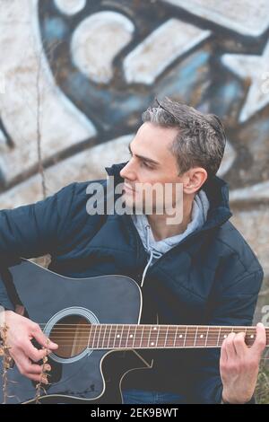A young white man, with gray hair, musician and artist, plays an elegant black guitar, and composes music outside, in the countryside, on a cloudy aft Stock Photo