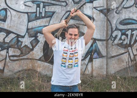 A young white man, with gray hair, musician and artist, holds a guitar behind him with his hands, while shouting in the countryside, on a cloudy after Stock Photo