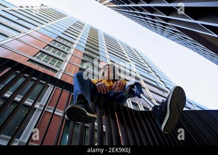 Disabled young man with leg prosthesis sitting on metal railing against skyscraper Stock Photo