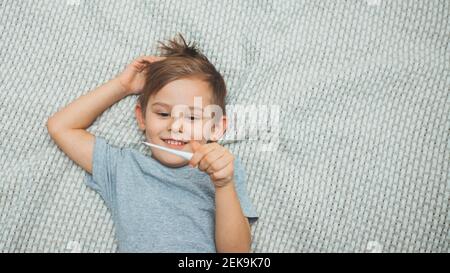 Little boy lying on the bed holding a thermometer in his hand and smiling. The temperature is normal. Health care. Coronavirus pandemic. Covid-19 Stock Photo