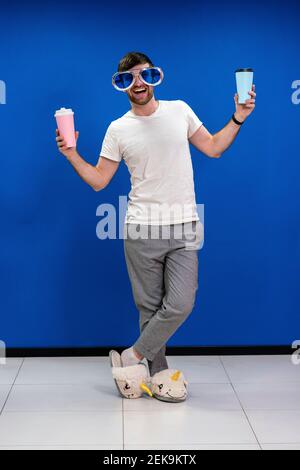 Happy businessman in unicorn slippers wearing novelty glasses while holding coffee cups against blue wall at work place Stock Photo