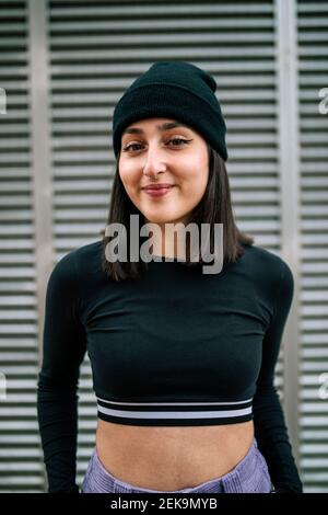 Smiling young woman with knit hat standing against metallic shutter Stock Photo