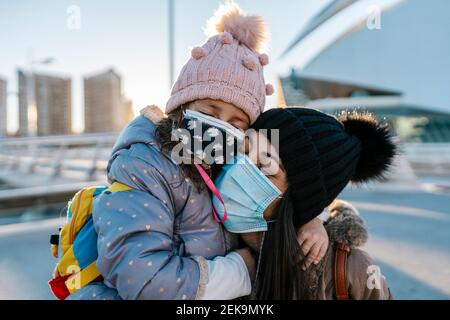 Mother and daughter wearing knit hat and face mask hugging while standing outdoors Stock Photo