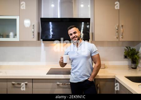 Handsome young man with hands in pockets holding coffee cup while standing in kitchen Stock Photo