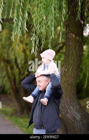 Grandfather carrying granddaughter on shoulders against tree in park Stock Photo