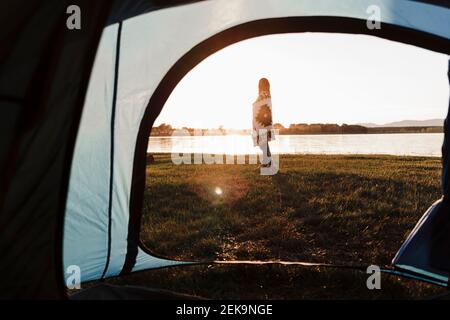 Woman wrapped in shawl standing on grassy land against lake seen through tent Stock Photo