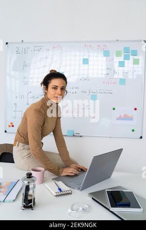 Young female student with laptop sitting on table by whiteboard Stock Photo