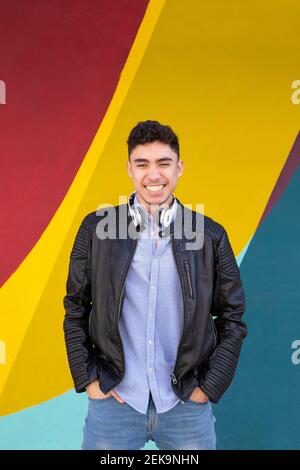 Happy man in leather jacket looking away while listening music by multi colored wall Stock Photo