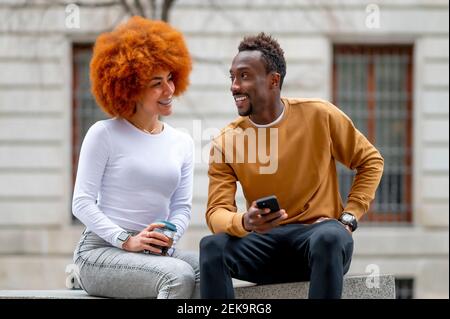 Smiling woman with disposable cup talking with man while sitting on bench Stock Photo