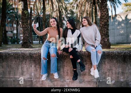 Smiling teenage girl taking selfie with friends while sitting on retaining wall in park Stock Photo