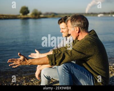 Father gesturing while talking with son by lake on sunny day Stock Photo