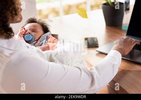 Mother holding sleeping baby while working on laptop at home Stock Photo