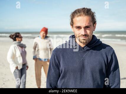 Portrait of young man wearing hooded shirt standing on beach with two young woman walking behind in background Stock Photo
