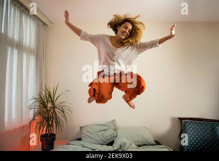 Cheerful young woman with arms raised jumping over bed in bedroom Stock Photo