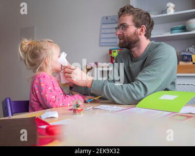Playful father holding paper on daughter's while sitting at table Stock Photo