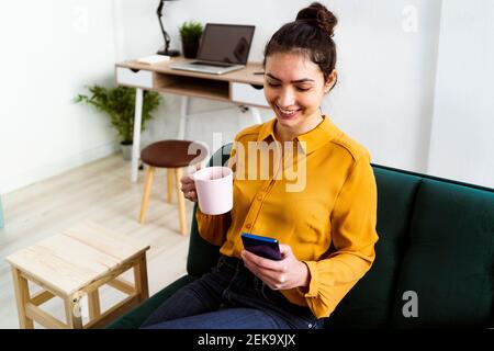 Smiling young woman drinking coffee while using mobile phone sitting at home Stock Photo