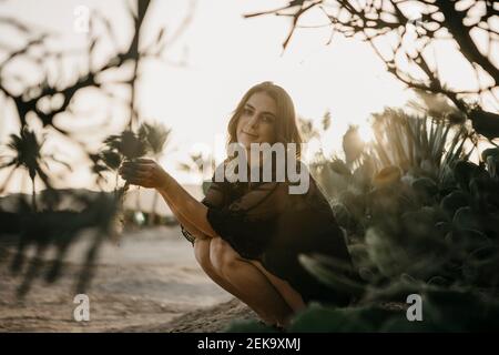 Smiling woman with sand crouching by cactus plant during sunset Stock Photo