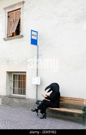 Female in crow costume using mobile phone while sitting on bench of bus stop Stock Photo