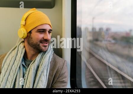 Smiling man wearing headphones looking through window while sitting in train Stock Photo