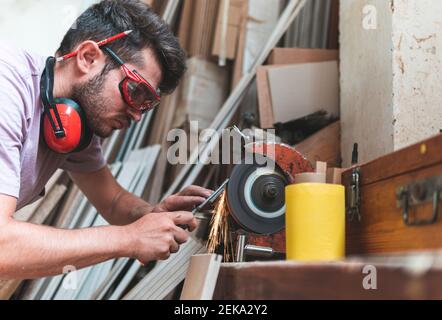 Concentrated male carpenter sharpening work tool on grinder in workshop Stock Photo