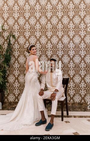 Beautiful bride standing by happy groom smiling while sitting on chair against wall Stock Photo