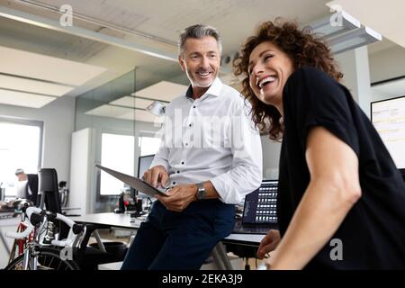 Smiling businessman with digital tablet looking at cheerful colleague while standing at open plan office Stock Photo
