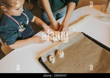 Mother holding cinnamon roll dough while daughter cutting with knife at table Stock Photo