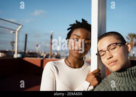Female friends with eyes closed leaning on pole Stock Photo