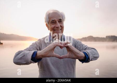 Smiling man making heart shape with hands while standing against lake Stock Photo