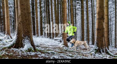 Young male sportsperson running with labrador during winter in forest Stock Photo
