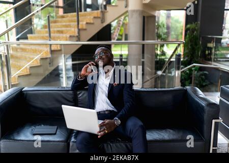 Handsome male businessman with laptop talking on phone call while sitting on sofa in hotel Stock Photo