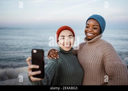 Smiling friends taking selfie through mobile phone while standing against sea Stock Photo