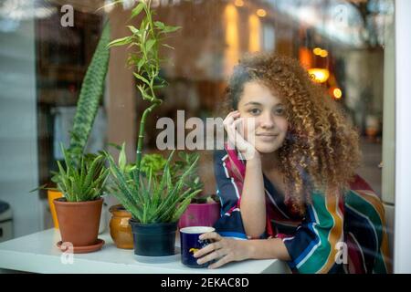 Smiling woman with coffee cup leaning by potted plant seen through glass at home Stock Photo