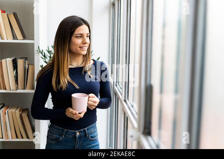 Smiling female freelancer with coffee cup looking through window while standing at home Stock Photo