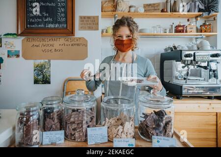 Female owner holding plate filled with chocolate while working in candy store during COVID-19 Stock Photo
