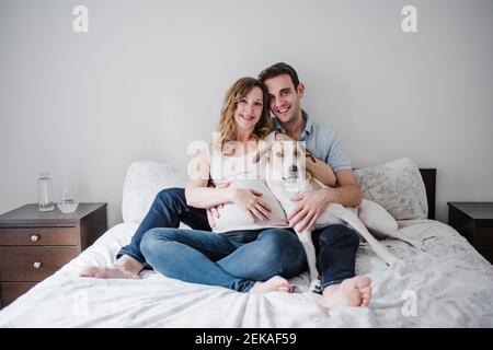 Smiling couple with dog relaxing on bed against wall at home Stock Photo