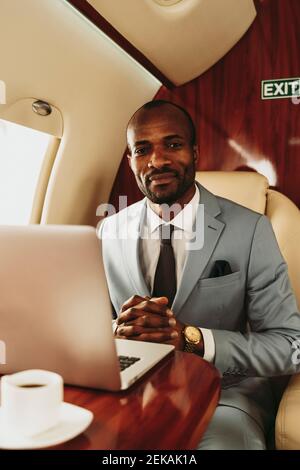 Smiling businessman with hands clasped by laptop in airplane Stock Photo