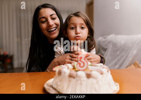 Girl blowing candle over birthday cake while sitting with mother at home Stock Photo