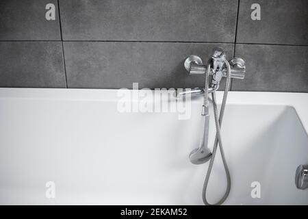 Chrome tap with a shower with a white bath in a bathroom with a grey tile. Stylish white bath with a silver tap. Minimalism in interior of bathroom. Stock Photo