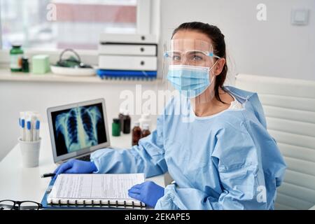 Female doctor wearing face shield while sitting at desk in medical clinic Stock Photo