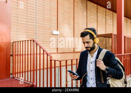 Mid adult businessman with backpack and headphones using mobile phone while standing by railing Stock Photo