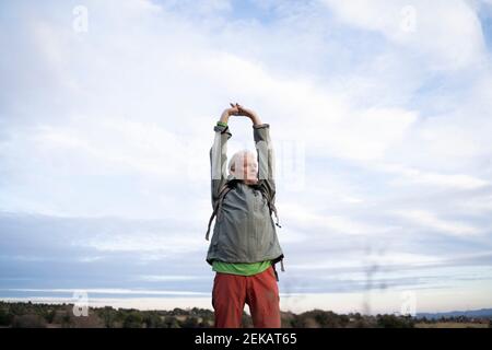 Senior male hiker stretching arms while standing against cloudy sky at countryside Stock Photo