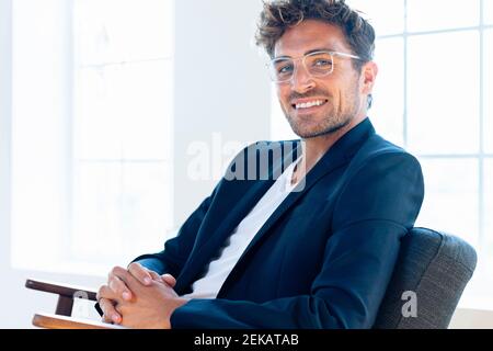 Confident businessman wearing eyeglasses sitting with hands clasped on armchair at home Stock Photo