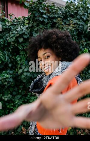 Smiling young woman gesturing while standing against green leaf wall